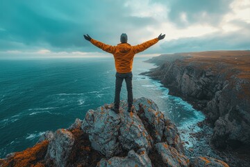 Wall Mural - A man stands on a rocky cliff overlooking the ocean, as the man seems to be celebrating the beauty of nature and the vastness of the ocean