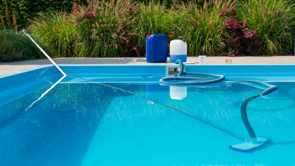 A clear blue pool with a submerged cleaning hose, chemical containers, and a water testing kit on the poolside, framed by lush greenery.