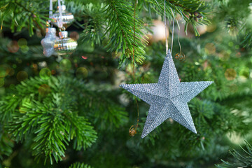 Silver Christmas star hanging on fir tree against blurred festive lights.