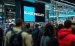 © lmot11 - A crowd of shoppers in a mall looks at a large screen with the text 'Black Friday'.  Consumerism concept. Sales and discounts.