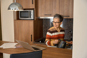 Smiling mature woman making tea while looking at laptop screen in hotel room