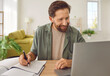 © Studio Romantic - Happy adult man studying online. Smiling bearded male student in casual shirt sitting at desk at home, using laptop computer, watching lecture, learning new information, taking notes in paper notebook