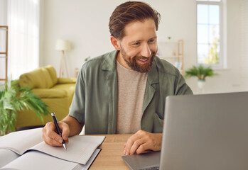 happy adult man studying online. smiling bearded male student in casual shirt sitting at desk at hom