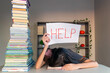 © Iryna - Overwhelmed student with head down holding a help sign. A student rests their head on a desk, exhausted from studying, while holding up a sign that says HELP.