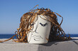 © Kira_Yan - Ceramic pot with hand-drawn woman face and dried plant leaves mimicking woven hair sits against blue sky
