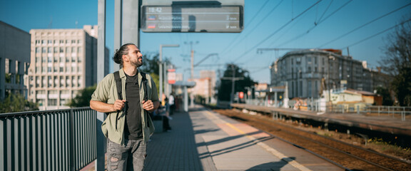 A man is waiting for a train on the platform. A young guy, a passenger with a backpack is standing on the platform waiting for the train