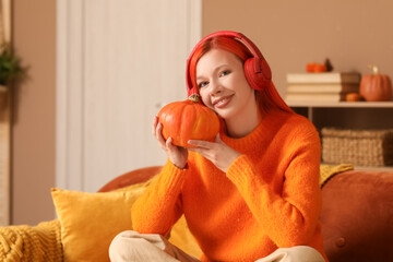 Poster - Beautiful young happy woman in headphones with pumpkin sitting on sofa at home