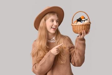 Sticker - Beautiful young happy woman pointing at wicker basket of Halloween pumpkins and gift box on grey background