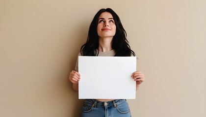 caucasian woman joyfully holding blank paper, standing against a beige background, looking upward in