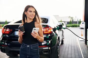 Wall Mural - With cup of drink and smartphone in hands. A young woman at a gas station with her car