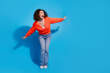 Wall Mural - Photo of wavy haired model woman wearing orange cardigan and jeans flying looking empty space curious isolated on blue color background