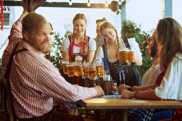 Two beautiful young women, waitress in traditional Bavarian clothes serving beer mugs to group of cheerful people. Concept of Oktoberfest, festival, party, brewery, traditions