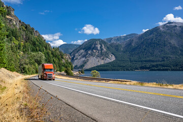 Wall Mural - Bright brown big rig semi truck transporting cargo in dry van semi trailer running on the winding highway road along the Columbia River with mountains range on the sides