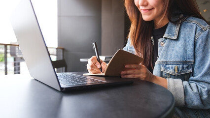 Wall Mural - Closeup image of a young woman writing on a notebook while working on laptop computer