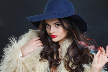 Wall Mural - Portrait of woman with red lips make-up and long wavy hair wearing blue hat against dark studio wall background