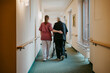 © Maskot - Rear view of female caregiver assisting senior woman walking with mobility walker in corridor