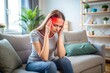 © Introvertia - Young woman suffers from migraine headache at home while sitting on comfortable couch holding head