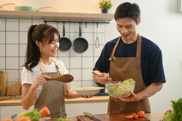 Wall Mural - Cheerful young couple preparing healthy vegetable salad in minimal kitchen