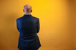 © karrastock - The image shows a man in a dark blue suit and a light blue shirt, standing with his back to the camera in front of a yellow wall.