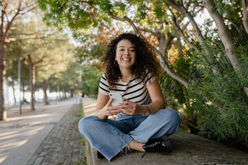 Wall Mural - pretty curly woman walking in city street in striped t-shirt, using smart phone