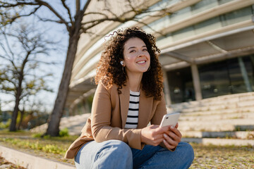 Wall Mural - pretty curly smiling woman walking in city street in stylish jacket, using smartphone