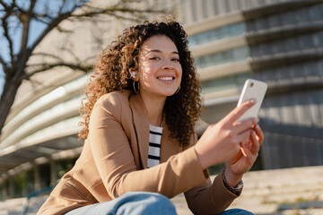 Wall Mural - pretty curly smiling woman walking in city street in stylish jacket, using smartphone