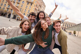 Tourist group of young excited friends together posing happy for photo in city square. Gen z people smiling with arms raised in air for holiday souvenir picture enjoying having fun in European town