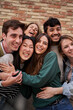 © CarlosBarquero - Vertical. Cheerful group of diverse young friends enjoying having fun together with brick wall in background. Multiracial happy generation z people posing with big smile hugging in community outdoors