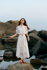 Poster - Woman in white dress embracing the ocean breeze on rocky shoreline