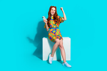 Wall Mural - Full size photo of pretty young girl sit cube hold device winning wear trendy colorful outfit isolated on aquamarine color background