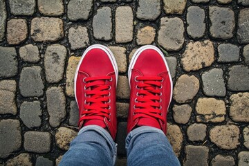 Wall Mural - Looking Down At Shoes. Top View of Man's Feet in Red Sneakers on Cobbled Road Background