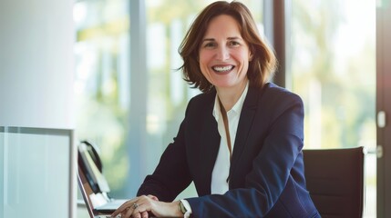 A businesswoman sitting by her laptop, dressed in a professional suit, smiling confidently with a modern office setting in the background.