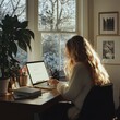 © Indi - female with long blond hair sitting at desk in front of laptop by window with view of trees and snow outside, daytime. remote working, freelancing lifestyle, online education, home office concept