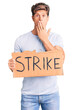 © Krakenimages.com - Young handsome man holding strike banner cardboard covering mouth with hand, shocked and afraid for mistake. surprised expression