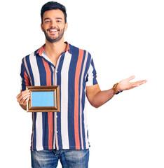 Young handsome hispanic man holding empty frame celebrating victory with happy smile and winner expression with raised hands