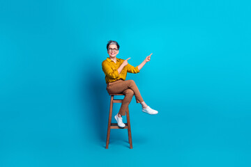 Wall Mural - Full length photo of charming positive woman wear blue blouse eyeglasses sitting chair pointing empty space isolated blue color background