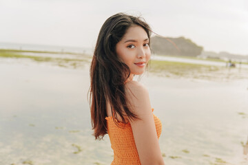 Joyful young Asian woman smiling at the camera from her shoulder with the ocean in the background.