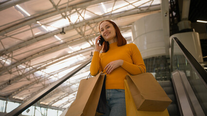 Joyful Caucasian woman female girl shopper buyer tourist shopaholic carrying shopping bags speaking talk smartphone mobile phone call discussing sales discount rides moving staircase inside shop mall