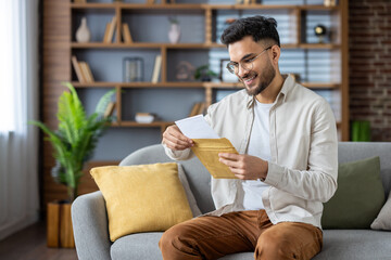 smiling man opening envelope with excitement while sitting on comfortable couch in modern home setti