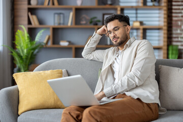 Poster - Thoughtful hispanic man working at home on laptop, sitting on comfortable couch in modern living room