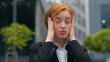 © Yuliia - Close up unhappy tired young Caucasian ginger woman gen z businesswoman girl female student formal suit holding head hands feeling anxiety pain sick disease discomfort healthcare outdoors city street
