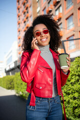 Poster - Vertical photo of pretty cheerful lady wear red leather jacket drinking beverage listen modern gadget outdoors city urban street