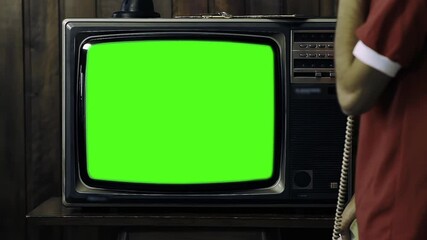 Wall Mural - man holding 80s phone while turning on old tv with green screen in a dimly lit room - you can replac