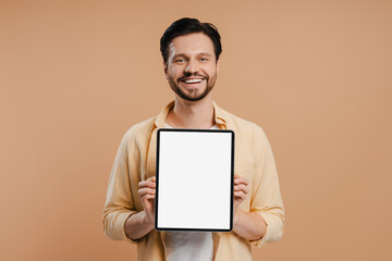 Happy man showing digital tablet with empty screen standing over beige background looking at camera