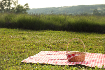 Picnic wicker basket with napkin and red checkered blanket on green grass outdoors