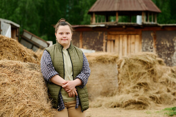 Portrait of smiling Caucasian woman with Down syndrome standing beside haystacks outdoors in rural area with rustic buildings visible in background