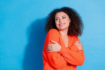 Wall Mural - Photo of young latin funny lady with curly hairstyle in stylish orange cardigan hugging herself looking dreamy isolated on blue color background