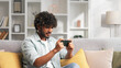 © YURII MASLAK - An Indian man is sitting on the sofa in his living room and playing an online game on his mobile phone, raising his hands up in victory celebration.