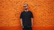 © Zhanna - A stylish young man wearing sunglasses stands confidently against a bright orange brick wall, hands in pockets. His dark outfit contrasts sharply with the vivid background, exuding a modern and cool
