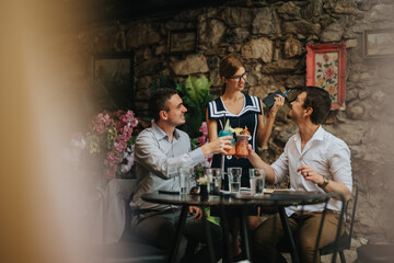 Poster - Three friends enjoying colorful drinks in a charming outdoor cafe with stone walls and floral decor, creating a warm and inviting atmosphere.